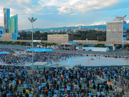 Elevated view of thousands of people gathered in Addis Ababa's Meskel Square, filling tiered seating and surrounding a large open plaza, with high-rise buildings and hills visible beneath a cloudy sky.