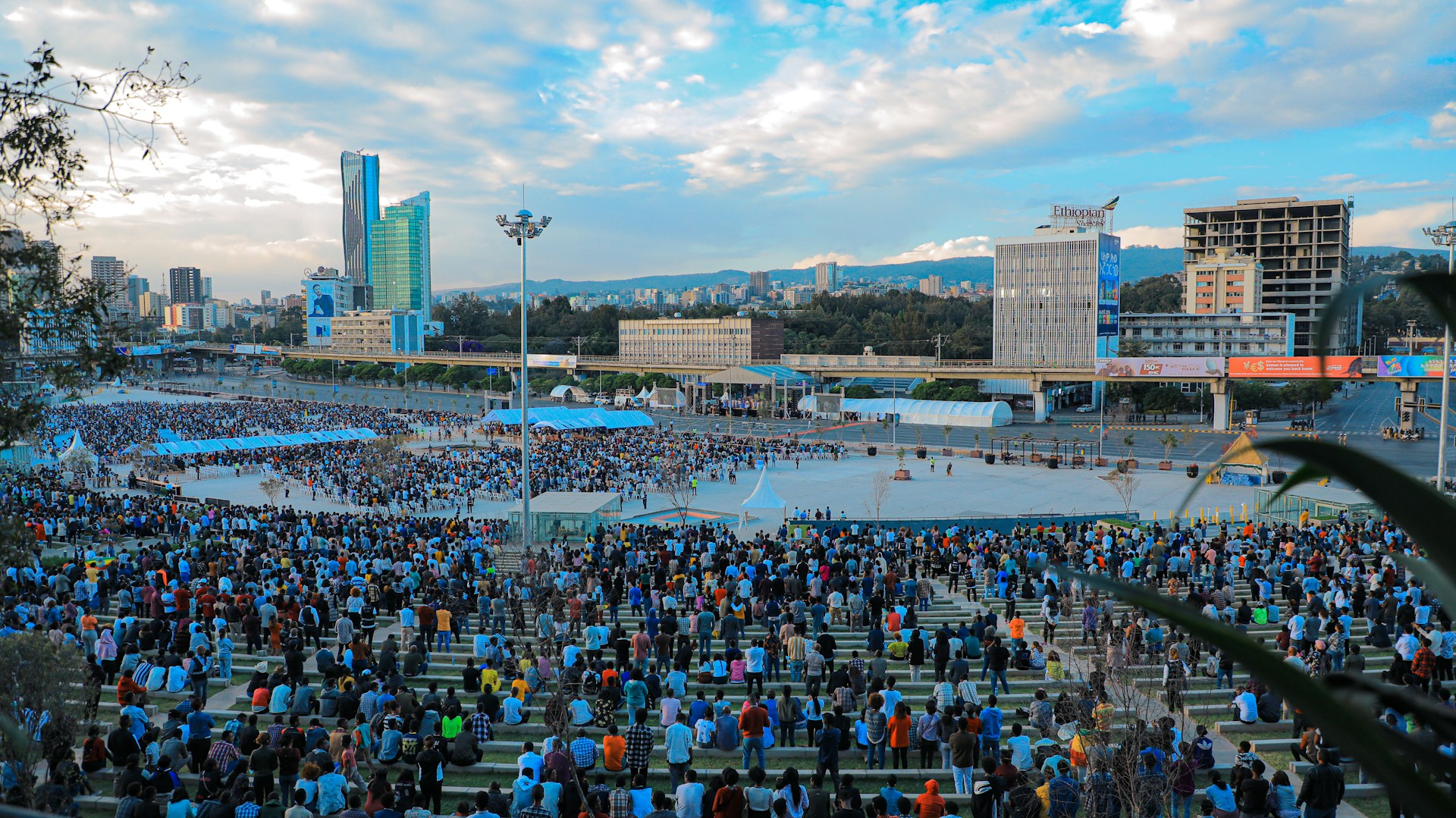 Elevated view of thousands of people gathered in Addis Ababa's Meskel Square, filling tiered seating and surrounding a large open plaza, with high-rise buildings and hills visible beneath a cloudy sky.