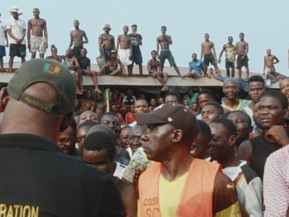 A large crowd of men gathers in the courtyard of New Bell Prison in Douala, with dozens more standing and sitting along the roof above them as a prison administrator faces the crowd.