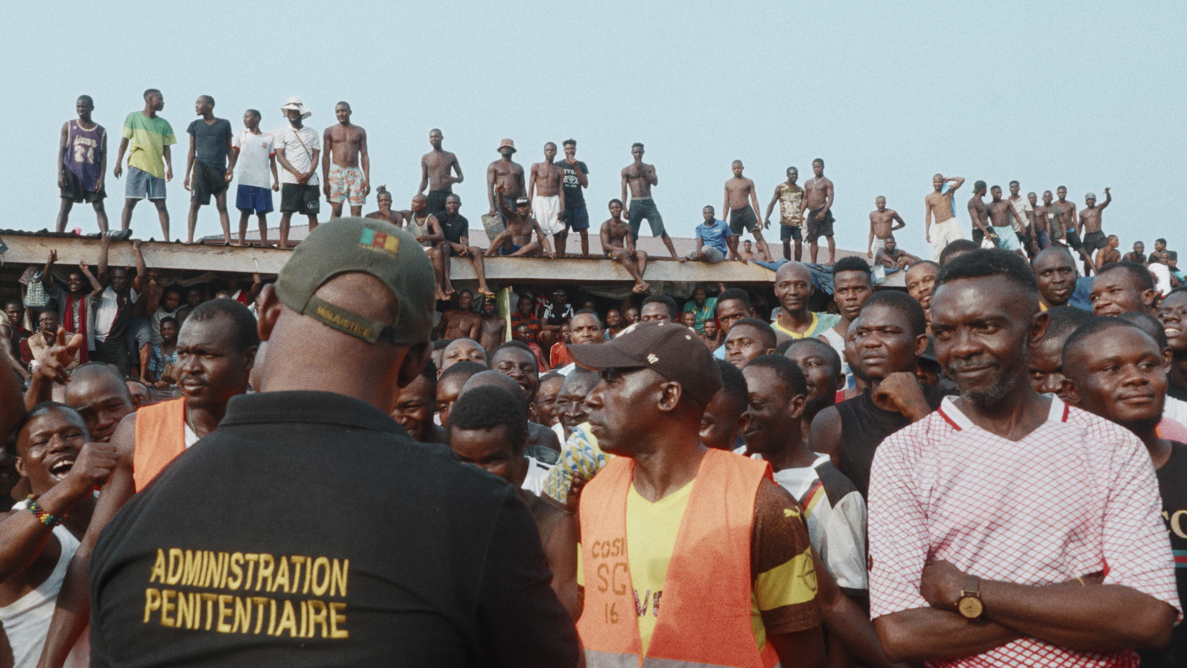 A large crowd of men gathers in the courtyard of New Bell Prison in Douala, with dozens more standing and sitting along the roof above them as a prison administrator faces the crowd.