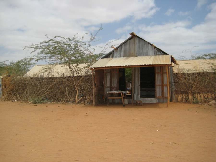 A store with a corrugated metal front, a peaked roof, and a wooden awning over a small table, set in a fence of woven acacia branches. Long, low corrugated-roof shelters stretch behind it on either side, above bare red earth