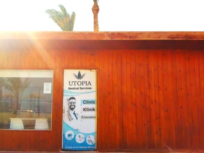 Exterior of Utopia Medical Services, a clinic housed in a reddish-brown wooden building, with a multilingual sign in English, German, and Russian on the entrance door. Palm trees rise above the roof as bright sunlight illuminates the building.