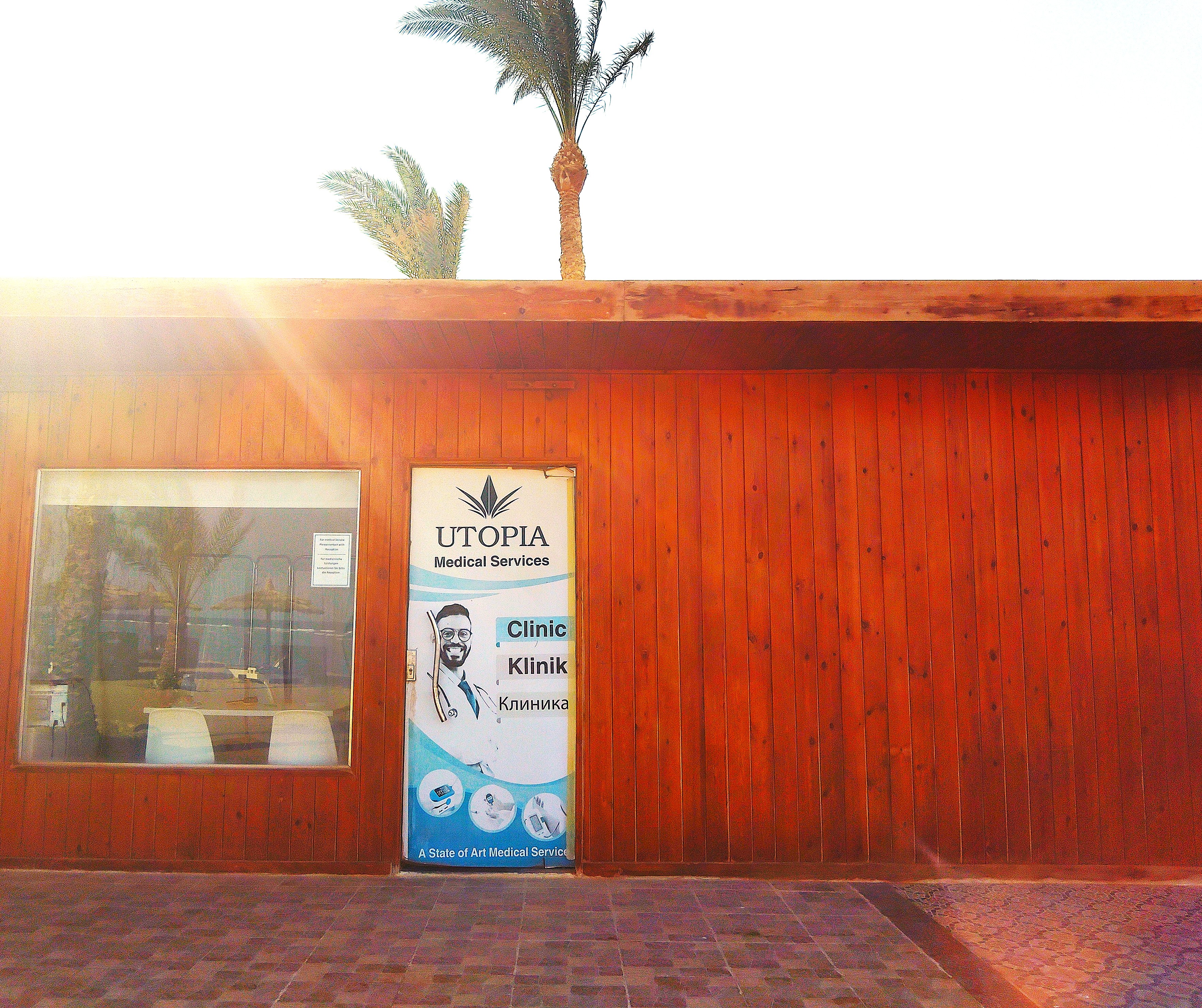 Exterior of Utopia Medical Services, a clinic housed in a reddish-brown wooden building, with a multilingual sign in English, German, and Russian on the entrance door. Palm trees rise above the roof as bright sunlight illuminates the building.