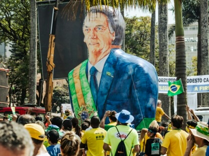A crowd of supporters dressed in Brazil’s green and yellow national colors gathers around a giant inflatable figure of former president Jair Bolsonaro wearing a presidential sash, with Brazilian flags waving beneath palm trees.