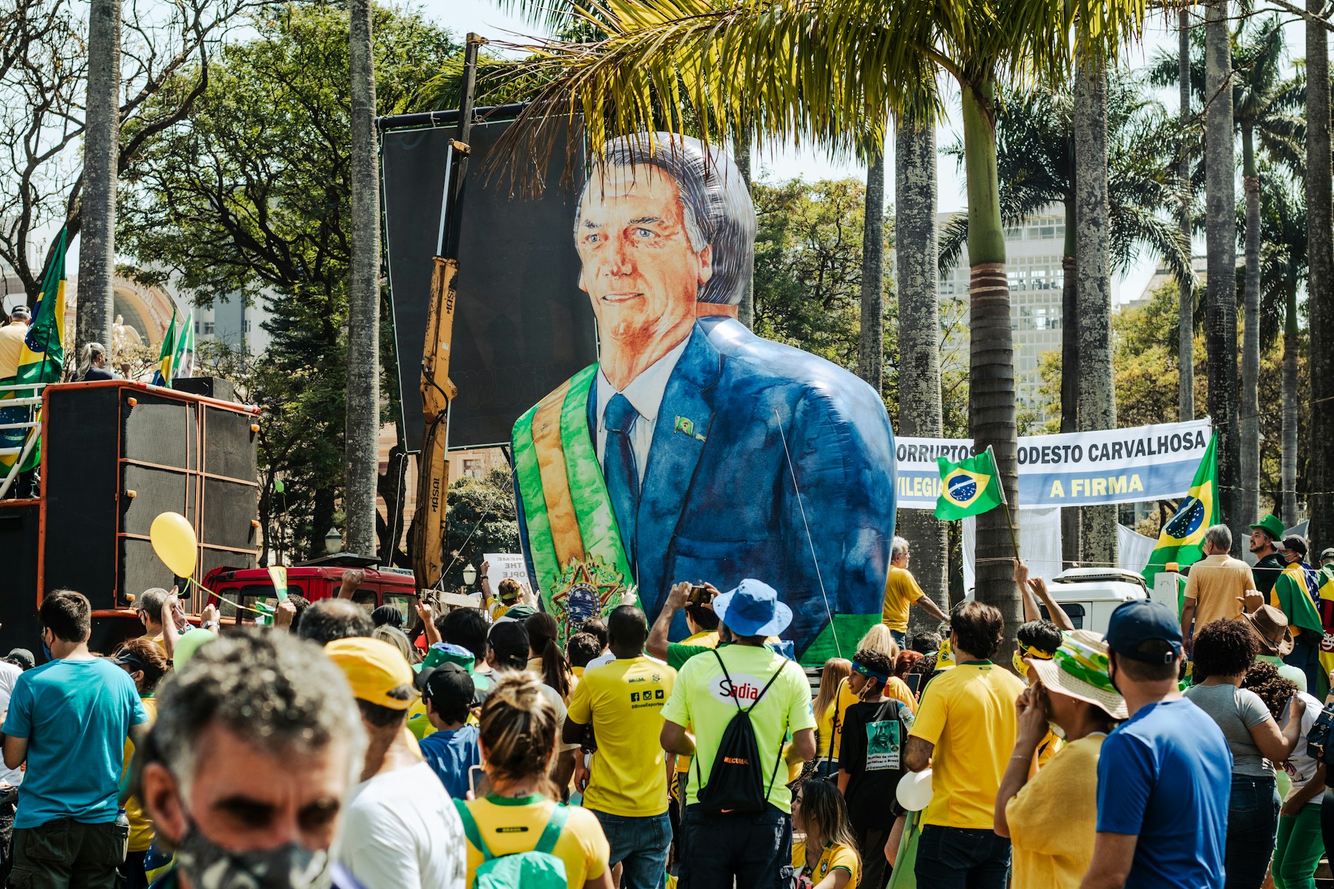 A crowd of supporters dressed in Brazil’s green and yellow national colors gathers around a giant inflatable figure of former president Jair Bolsonaro wearing a presidential sash, with Brazilian flags waving beneath palm trees.
