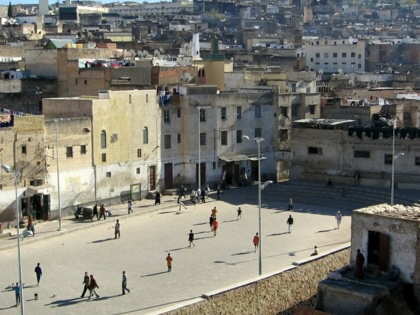 Elevated view of a large public square in a densely built Moroccan city, where children play football and pedestrians cross the open space, surrounded by weathered buildings stretching up a hillside.