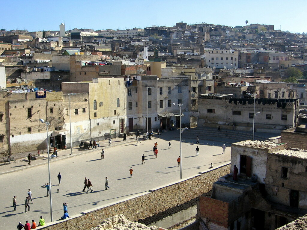 Elevated view of a large public square in a densely built Moroccan city, where children play football and pedestrians cross the open space, surrounded by weathered buildings stretching up a hillside.