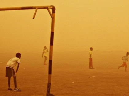 Children play football on a dusty, open field in Sudan as a sandstorm turns the sky deep orange. One child stands beneath a makeshift goalpost while others run across the pitch, partially obscured by blowing dust.
