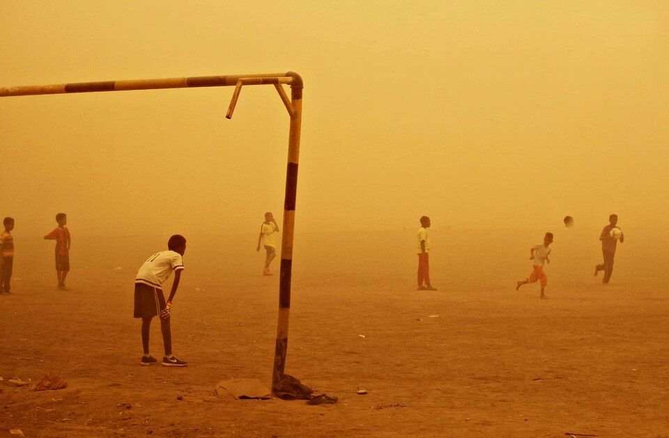 Children play football on a dusty, open field in Sudan as a sandstorm turns the sky deep orange. One child stands beneath a makeshift goalpost while others run across the pitch, partially obscured by blowing dust.