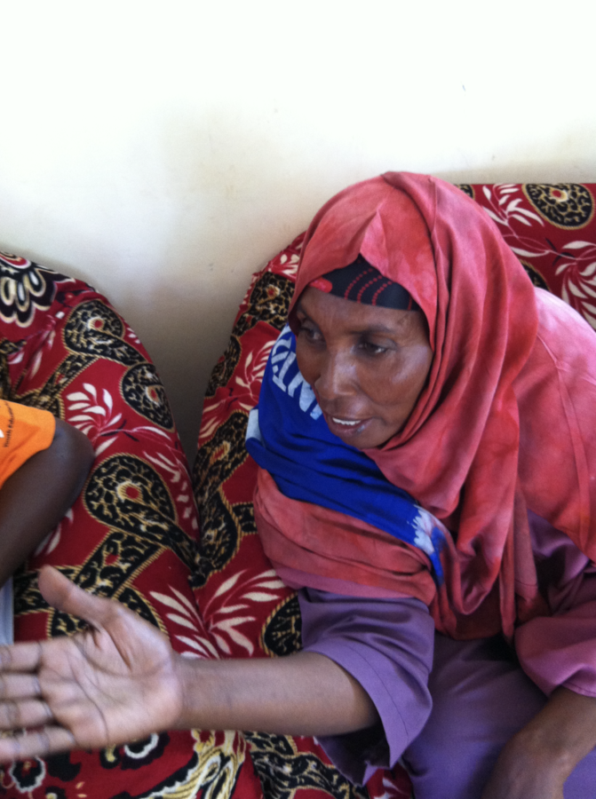 Isnina Ali Rage, wearing a red headscarf over a black headband and a blue scarf, sits on a sofa covered in red patterned fabric. She is mid-conversation, one hand stretched out with the palm open.