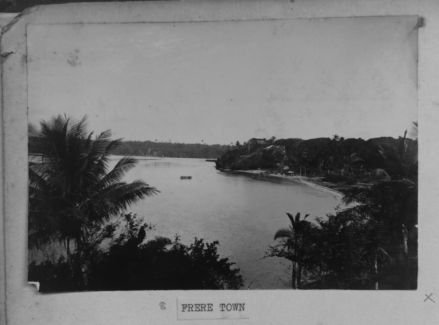 A black-and-white archival photograph, mounted in an album and labeled “Frere Town,” looking down through palm trees onto a calm bay. A boat with several people in it crosses the water toward a curved beach backed by dense trees and a few buildings.
