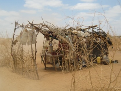 A small open-sided shop built from branches and thorny acacia, with plastic sheeting and cardboard across its roof. Goods are laid out on a wooden table inside, yellow jerrycans sit beside it, and dust blows across the flat, sandy ground under a partly cloudy sky.
