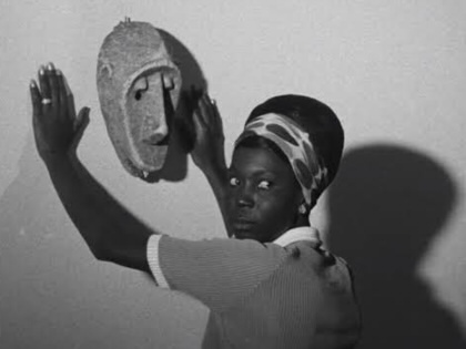 Black-and-white still from Black Girl showing Diouana, wearing a patterned headscarf, standing with her hands raised toward an African mask mounted on a wall, her face turned toward the camera.
