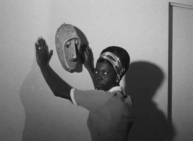 Black-and-white still from Black Girl showing Diouana, wearing a patterned headscarf, standing with her hands raised toward an African mask mounted on a wall, her face turned toward the camera.