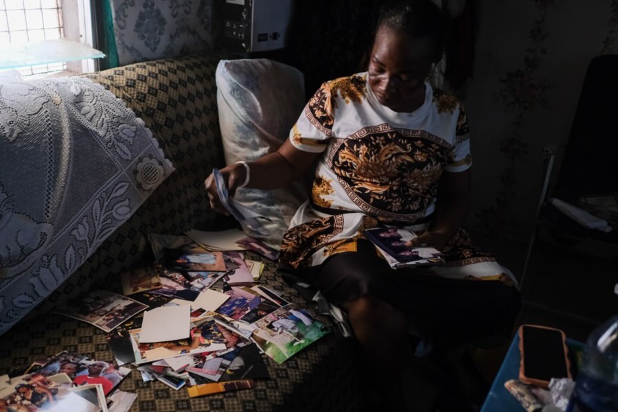 A women sifts through photographs and clippings, sitting on a couch in a darkly lit room.