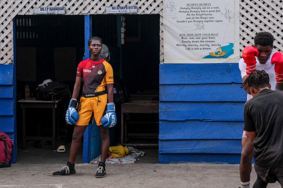 A boxer stands outside of a boxing gym, wearing blue boxing gloves.