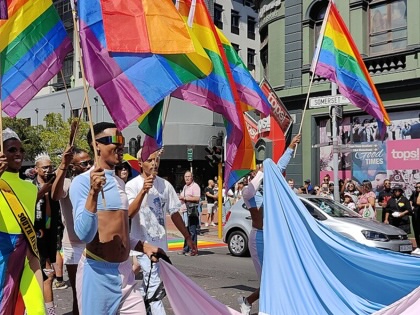 Participants in the Cape Town Pride parade walk along Somerset Road carrying rainbow flags and flowing pastel banners, while spectators watch from the roadside.