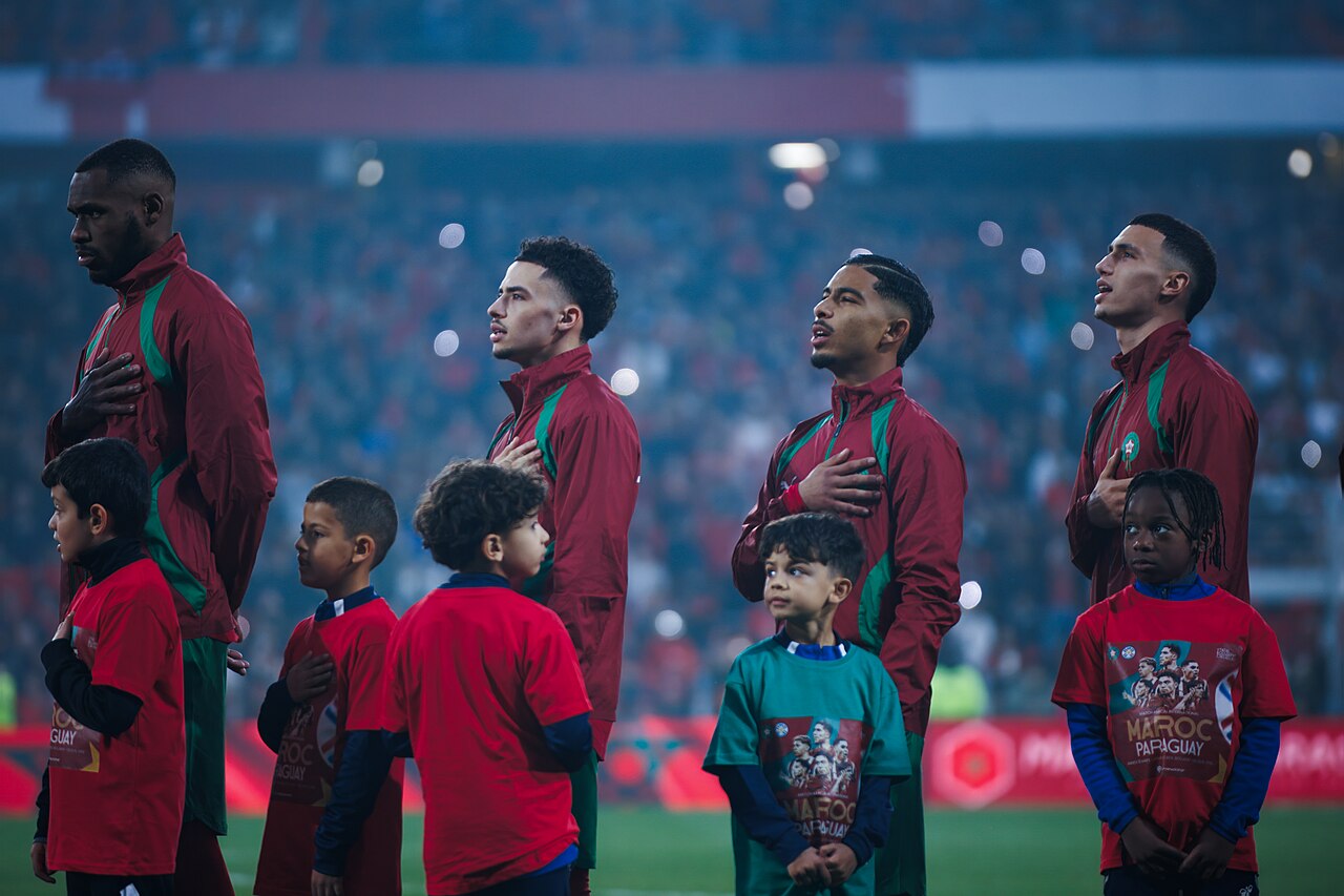 Moroccan footballers stand with their hands over their hearts during the national anthem before a match against Paraguay, accompanied by child mascots on the pitch.