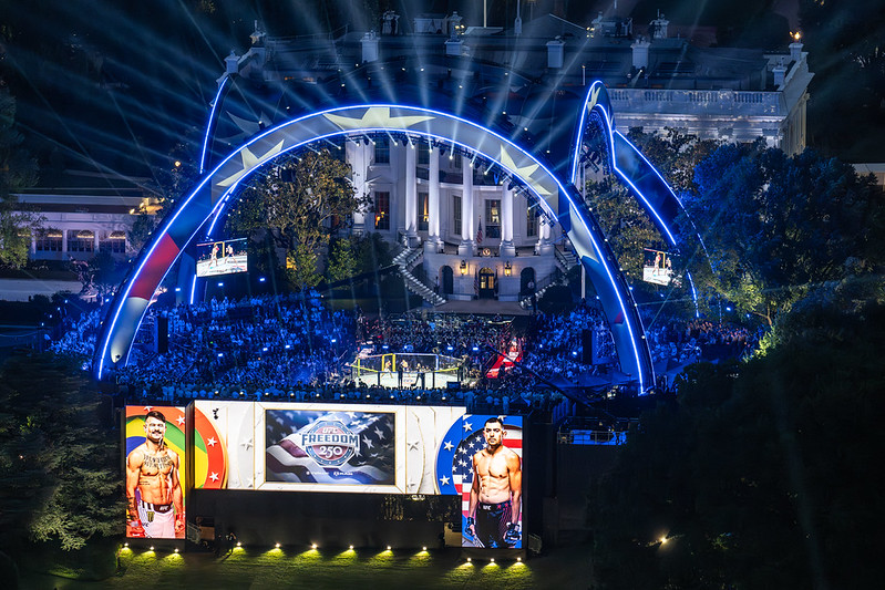 Nighttime view of UFC Freedom 250 on the Ellipse in Washington, D.C., with the White House illuminated in the background as spectators surround an outdoor octagonal cage.