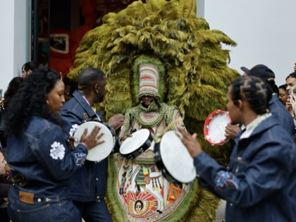 Performers play hand drums around a masked figure wearing an elaborate green feathered costume during a crowded cultural procession, as onlookers gather and take photographs outside a building.