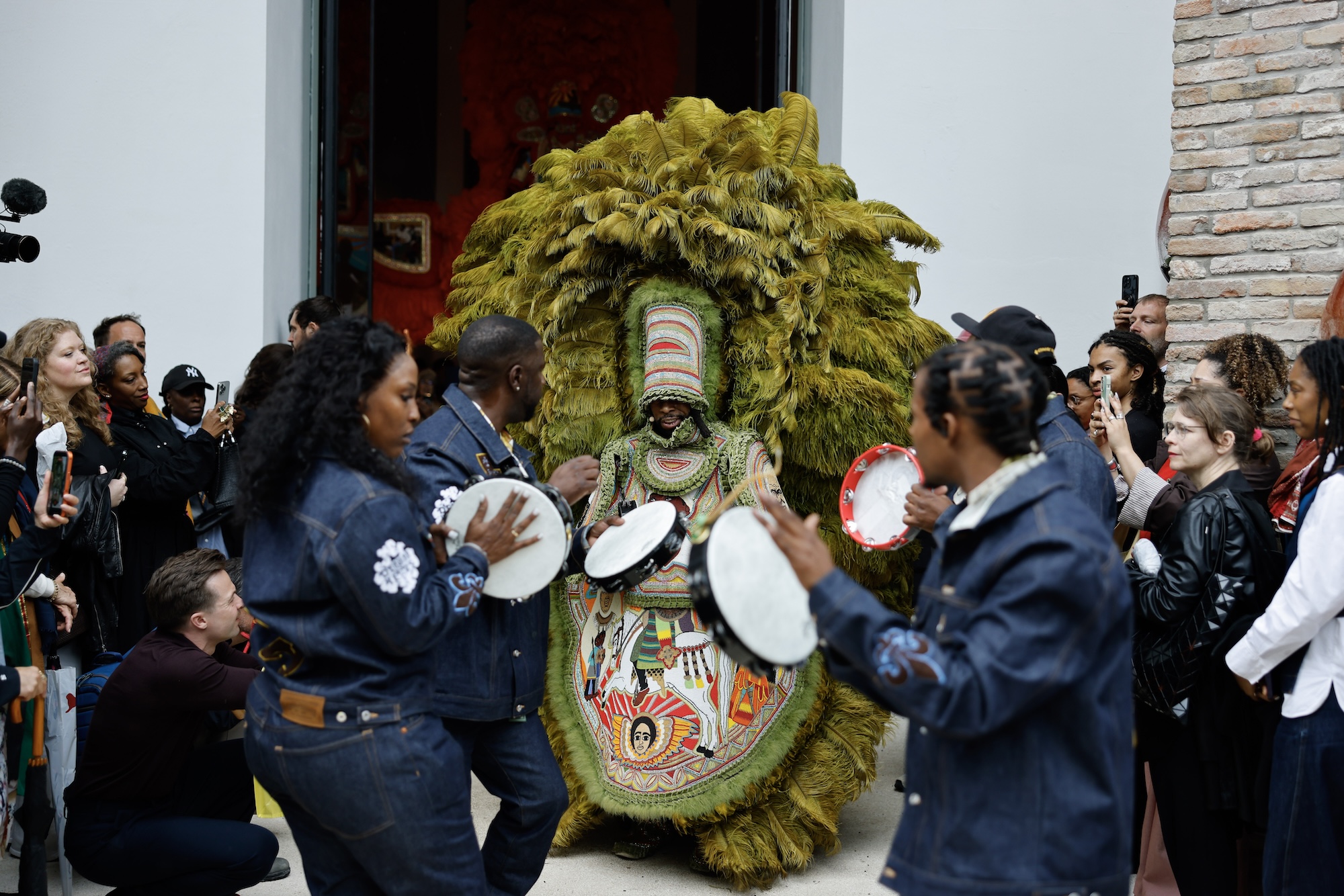 Performers play hand drums around a masked figure wearing an elaborate green feathered costume during a crowded cultural procession, as onlookers gather and take photographs outside a building.