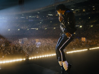 On a stadium stage at night, a singer in a black jacket and white socks stands in profile with a microphone, as a packed crowd stretches into the background under floodlights.