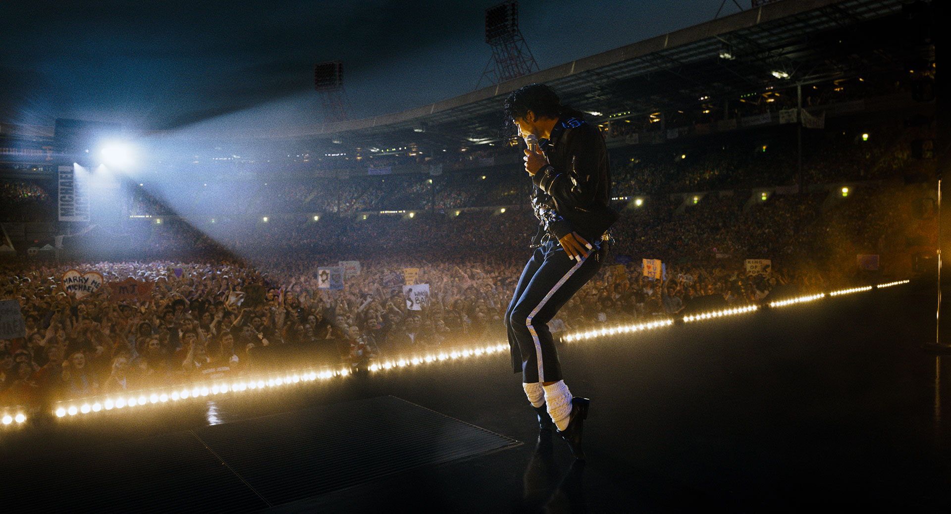 On a stadium stage at night, a singer in a black jacket and white socks stands in profile with a microphone, as a packed crowd stretches into the background under floodlights.