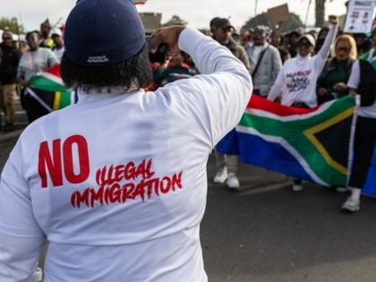A person wearing a shirt reading “No Illegal Immigration” stands with their back to the camera facing a crowd of demonstrators carrying South African flags during a March and March protest in Bellville, Cape Town.