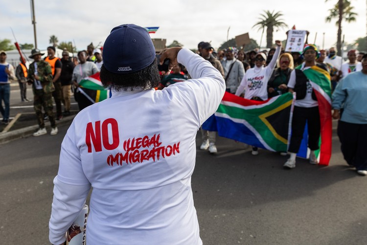 A person wearing a shirt reading “No Illegal Immigration” stands with their back to the camera facing a crowd of demonstrators carrying South African flags during a March and March protest in Bellville, Cape Town.
