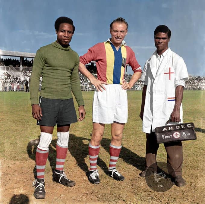 Stanley Matthews stands in a Hearts of Oak jersey between goalkeeper Lamptey Mills and a club physiotherapist on a football pitch in Accra during his 1957 visit to Ghana.