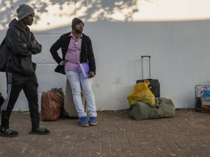 Three people stand and sit beside suitcases and bags on a pavement against a white wall, appearing to wait outside with their belongings.