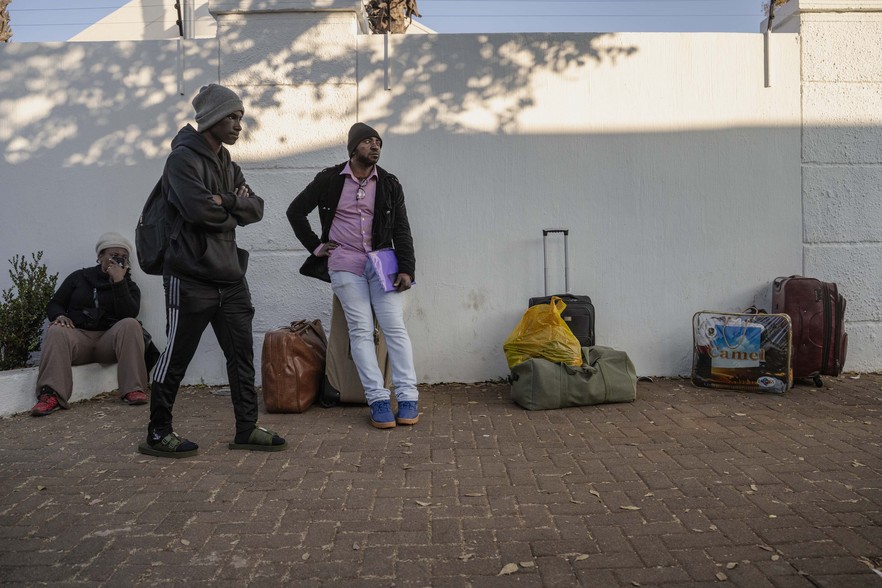 Three people stand and sit beside suitcases and bags on a pavement against a white wall, appearing to wait outside with their belongings.