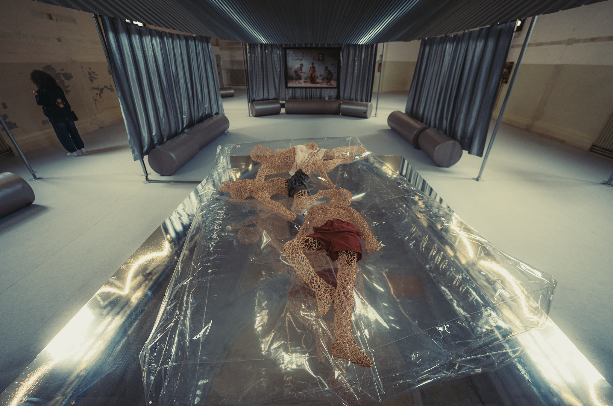 Installation view of a contemporary art exhibition featuring woven human-like figures reclining on a reflective platform covered in clear plastic, surrounded by curtained viewing spaces and seating within a large gallery.