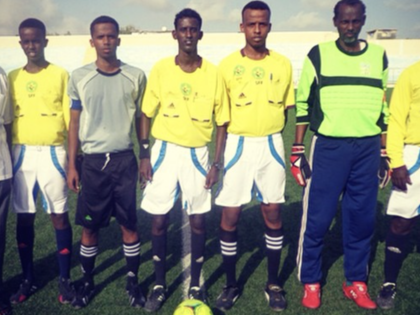 Match officials and team staff pose on a football field before a local match in Somalia, with referees in yellow kits standing around a ball at midfield.