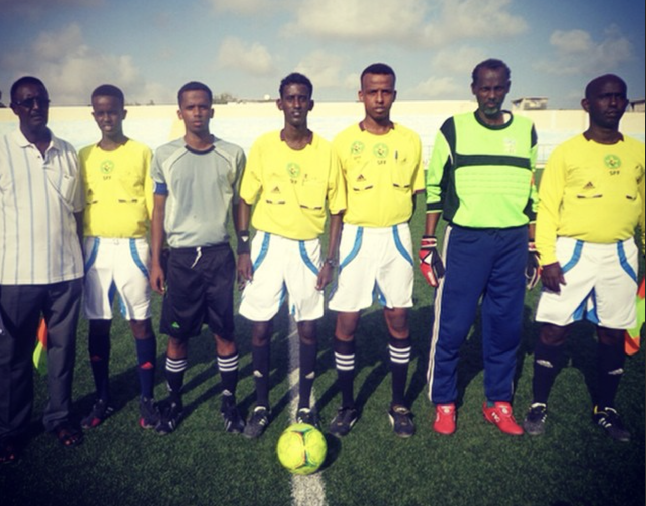 Match officials and team staff pose on a football field before a local match in Somalia, with referees in yellow kits standing around a ball at midfield.
