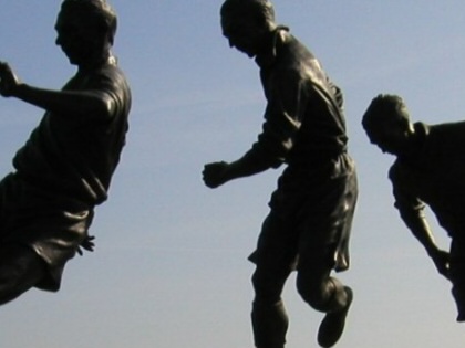 lhouetted statue of footballer Stanley Matthews in three poses, showing him dribbling a ball against a bright sky outside a stadium.