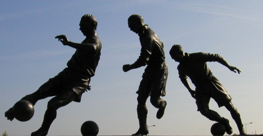 lhouetted statue of footballer Stanley Matthews in three poses, showing him dribbling a ball against a bright sky outside a stadium.