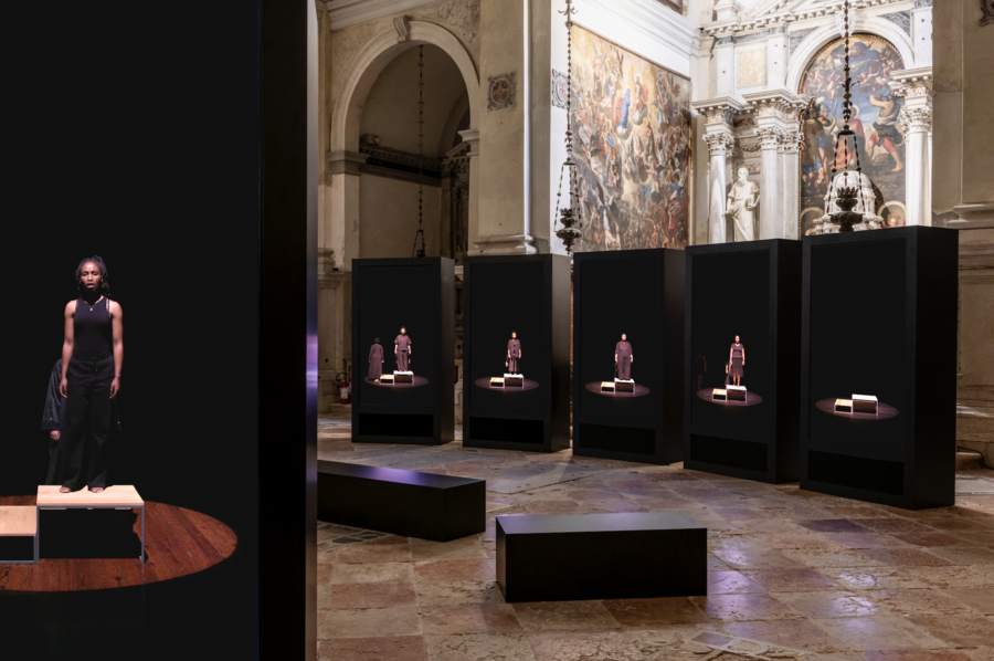 Installation view of a contemporary video artwork displayed inside a historic church, with multiple black screens showing life-sized figures standing on small platforms against dark backgrounds, framed by ornate arches, sculptures, and religious paintings.