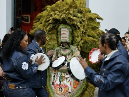 Performers play hand drums around a masked figure wearing an elaborate green feathered costume during a crowded cultural procession, as onlookers gather and take photographs outside a building.
