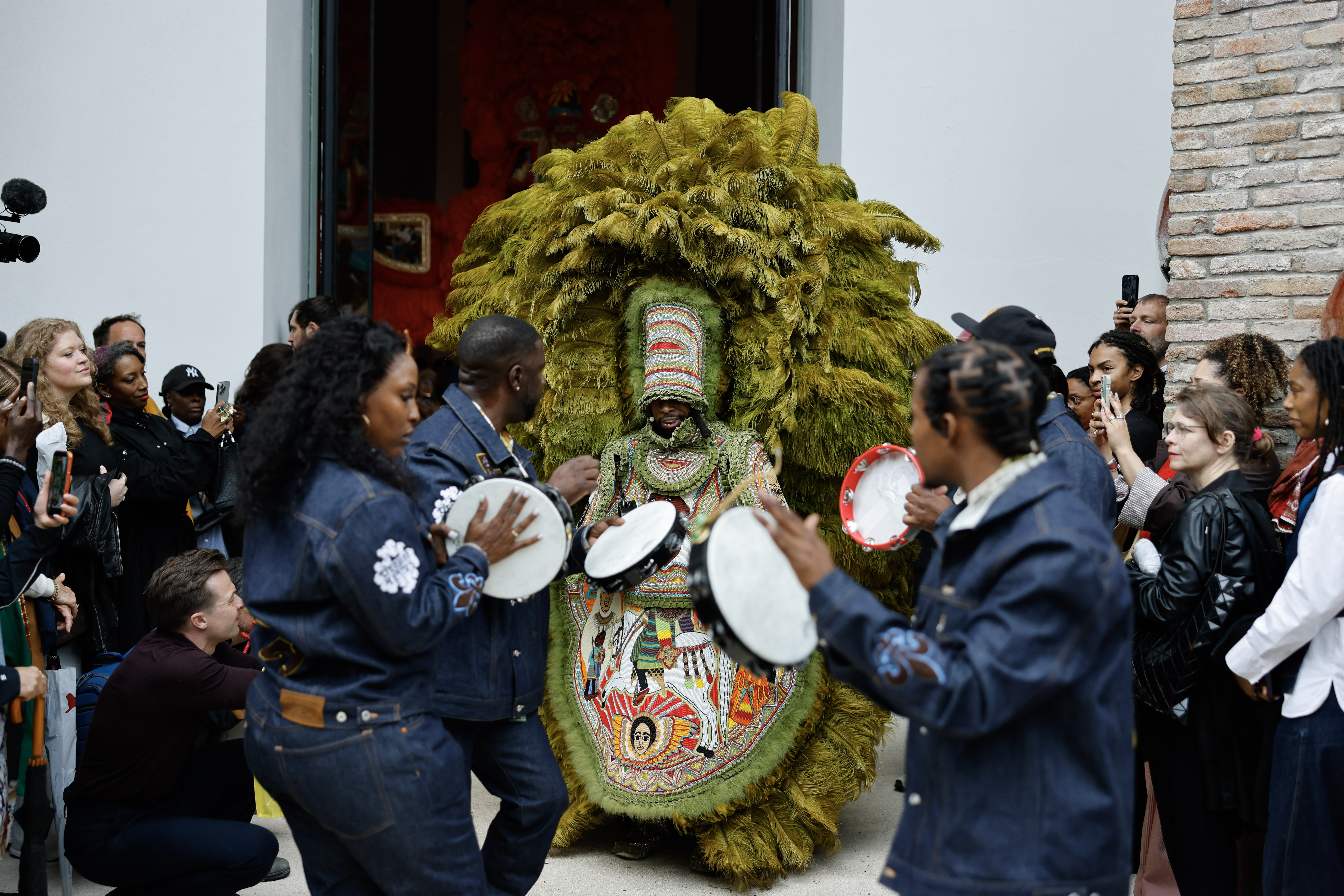 Performers play hand drums around a masked figure wearing an elaborate green feathered costume during a crowded cultural procession, as onlookers gather and take photographs outside a building.