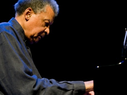 Abdullah Ibrahim sits at a piano on a dark stage, illuminated by spotlight as he performs during a concert.