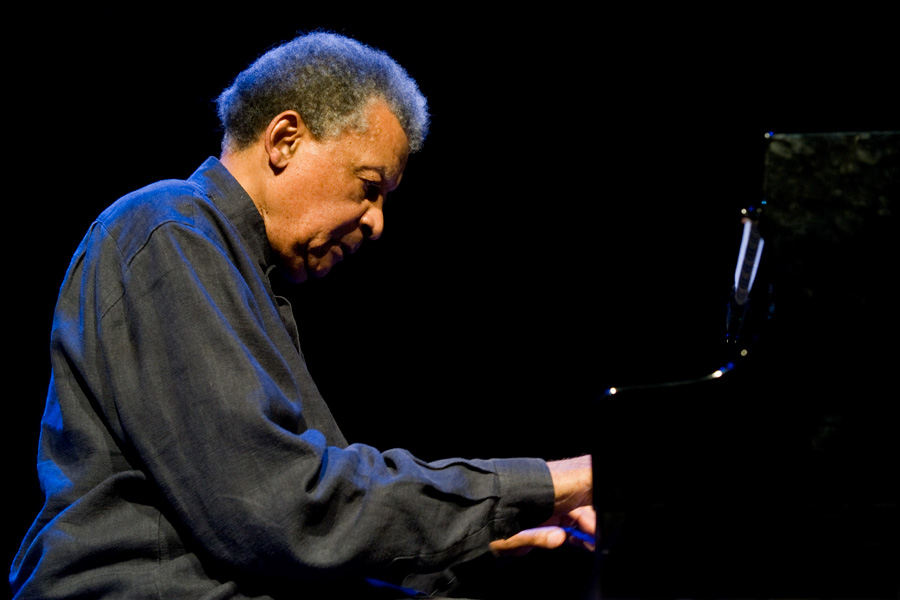 Abdullah Ibrahim sits at a piano on a dark stage, illuminated by spotlight as he performs during a concert.