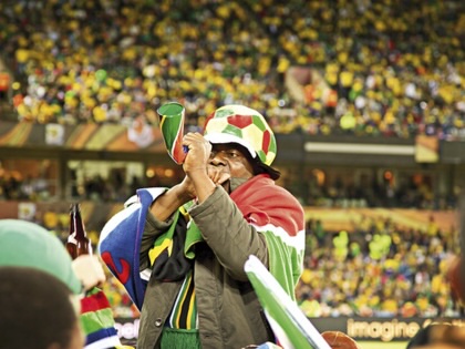 South African football supporter wearing a multicoloured hat and flag blows a vuvuzela in a crowded stadium during the 2010 FIFA World Cup.