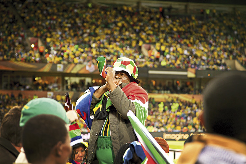 South African football supporter wearing a multicoloured hat and flag blows a vuvuzela in a crowded stadium during the 2010 FIFA World Cup.