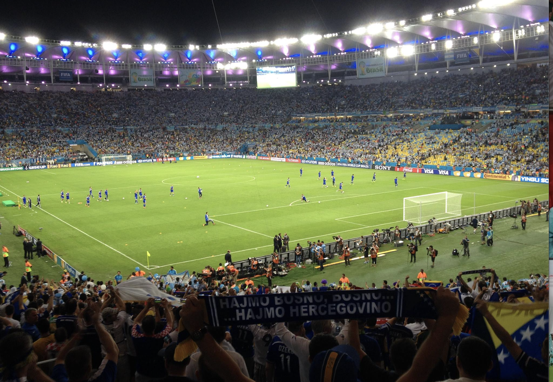 Players warming up on a football pitch in a packed stadium at nighttime.