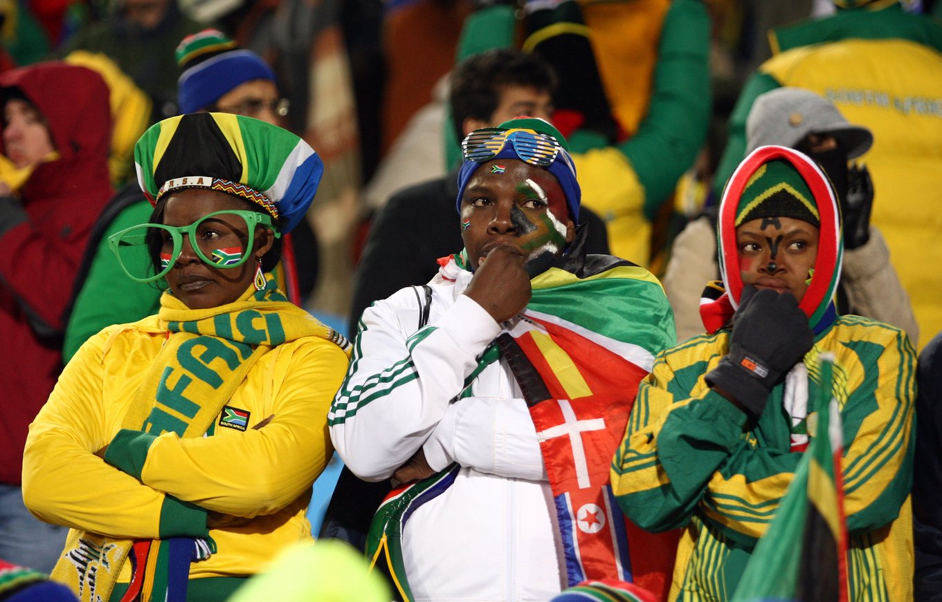 Three South African football supporters dressed in national colours sit silently in the stands, appearing dejected during a 2010 FIFA World Cup match against Uruguay.