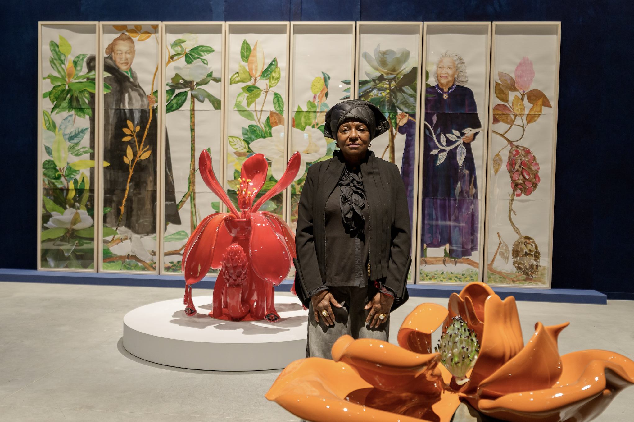 Alt text:
A woman dressed in black stands in a gallery between large, brightly colored floral sculptures, with a multi-panel artwork depicting plants and two women in the background.