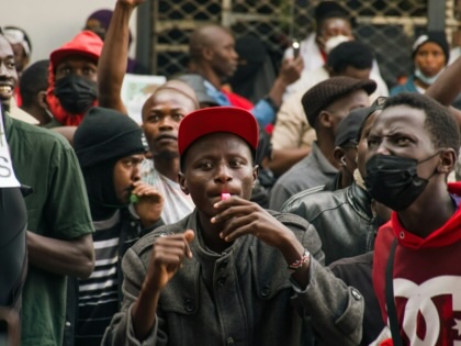 A crowd of protesters gathers in a city street, with several people raising clenched fists. In the foreground, a person in a red cap blows a whistle while others chant, some wearing face masks and holding placards.