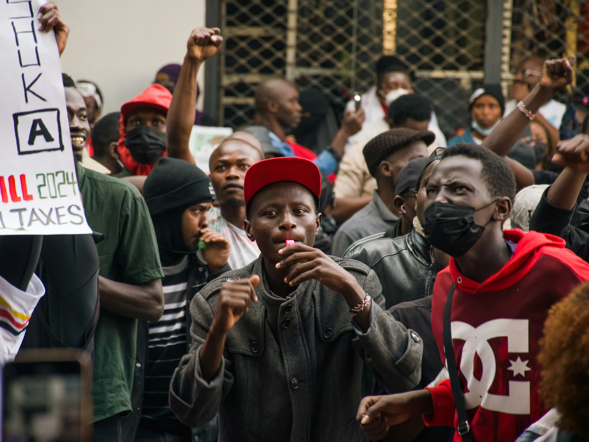 A crowd of protesters gathers in a city street, with several people raising clenched fists. In the foreground, a person in a red cap blows a whistle while others chant, some wearing face masks and holding placards.