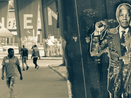 A weathered portrait of Nelson Mandela raising his fist is pasted to a concrete pillar beneath an overpass, while pedestrians walk along the street in the background.